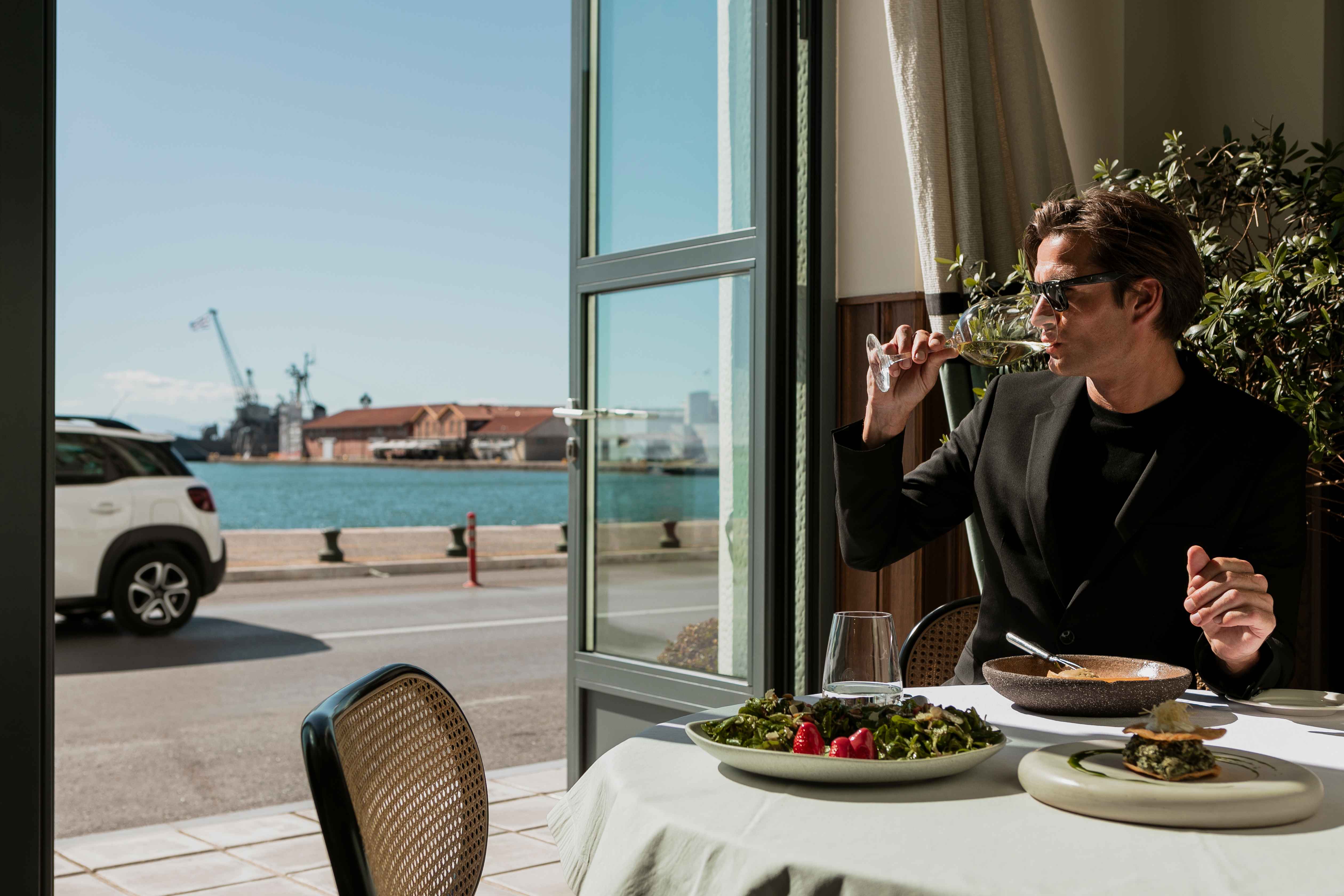 
Man in black suit enjoying lunch with wine by the window overlooking Thessaloniki port at ON Residence